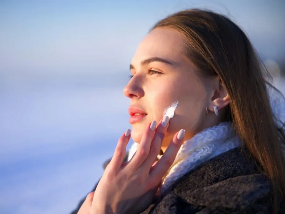 Woman applying moisturizer to her face while wearing a heavy winter coat to protect her skin from cold, dry weather