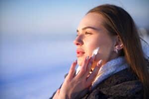 Woman applying moisturizer to her face while wearing a heavy winter coat to protect her skin from cold, dry weather